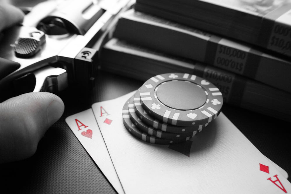 hand holding a gun next to stacks of money, poker cards and chips